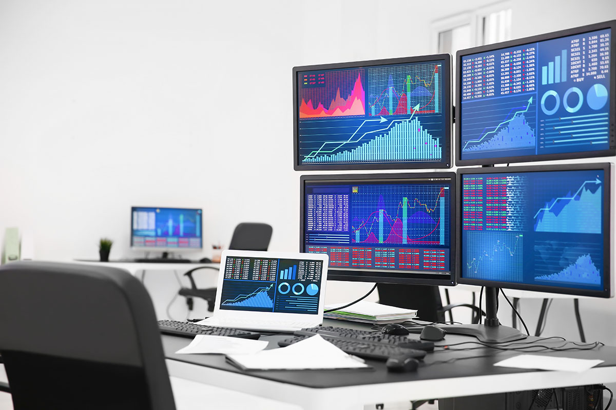 Four monitors and a laptop displaying financial charts, bar graphs and market data on a desk in a bright, empty office