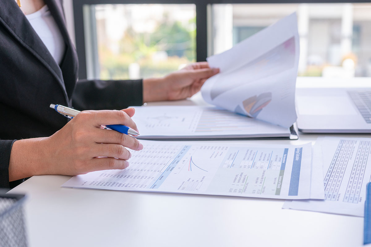 A professional in a dark suit flips through printed financial reports with charts while holding a pen over a spreadsheet summary on a white desk beside an open laptop