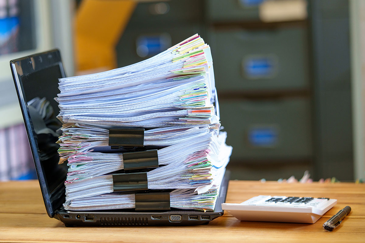A tall stack of paper documents held with black binder clips piled on an open laptop, beside a white calculator and a pen on a wooden desk, with filing cabinets blurred in the background