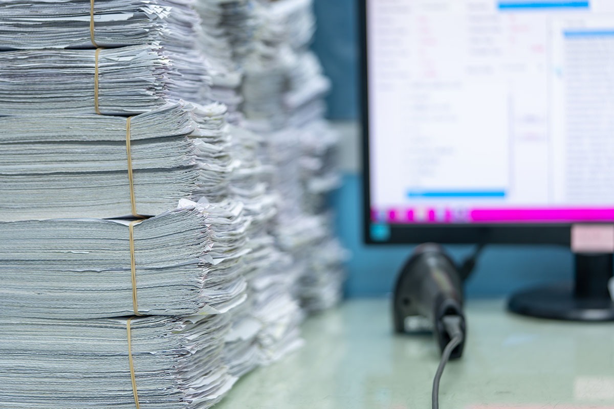 Tall stacks of paper documents bound with rubber bands on an office desk beside a desktop monitor showing a software window and a handheld barcode scanner