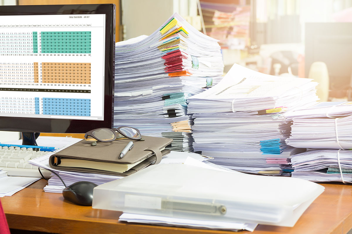 An office desk piled with tall stacks of tabbed paper documents beside a monitor showing a color-coded spreadsheet, with reading glasses and a pen resting on a leather notebook