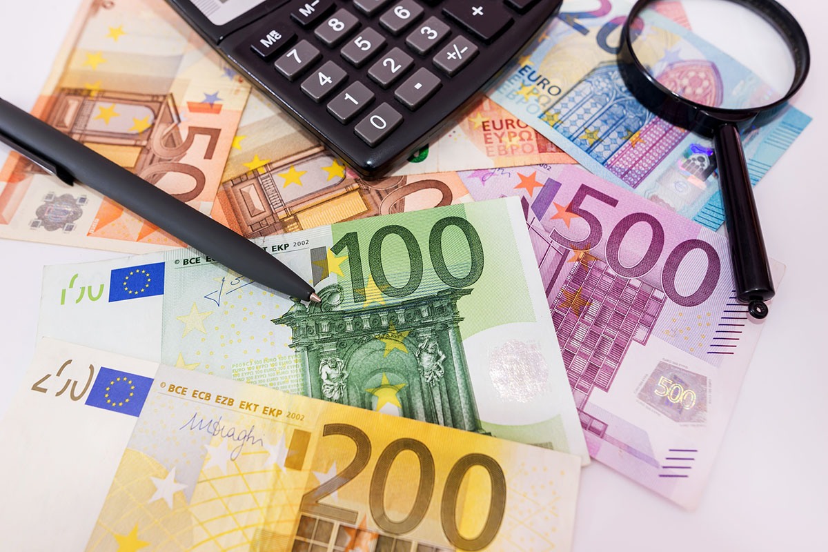 Euro banknotes in 50, 100, 200 and 500 denominations spread on a white desk with a black calculator, a pen and a magnifying glass