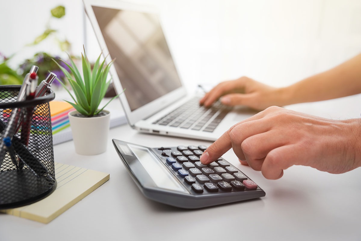 Close-up of a hand tapping keys on a black desktop calculator while the other hand types on a silver laptop, with a small potted succulent, a pen cup and a notepad on a white desk