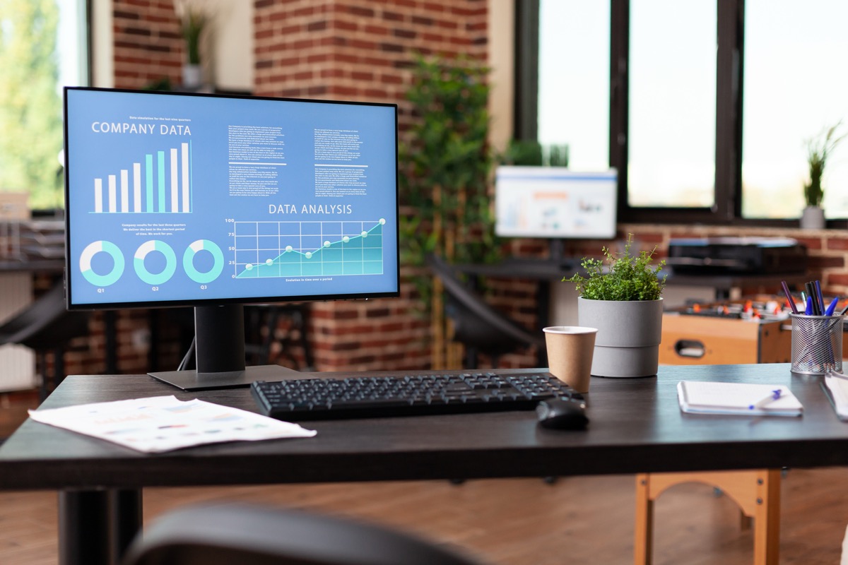A desk monitor showing company data bar charts, donut charts and a line graph beside a keyboard, paper reports and a coffee cup in a bright brick-walled office