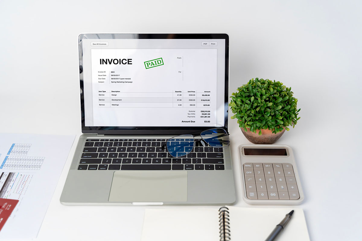 A laptop on a white desk displaying a digital invoice marked paid, beside a calculator, reading glasses, a potted plant and printed financial reports
