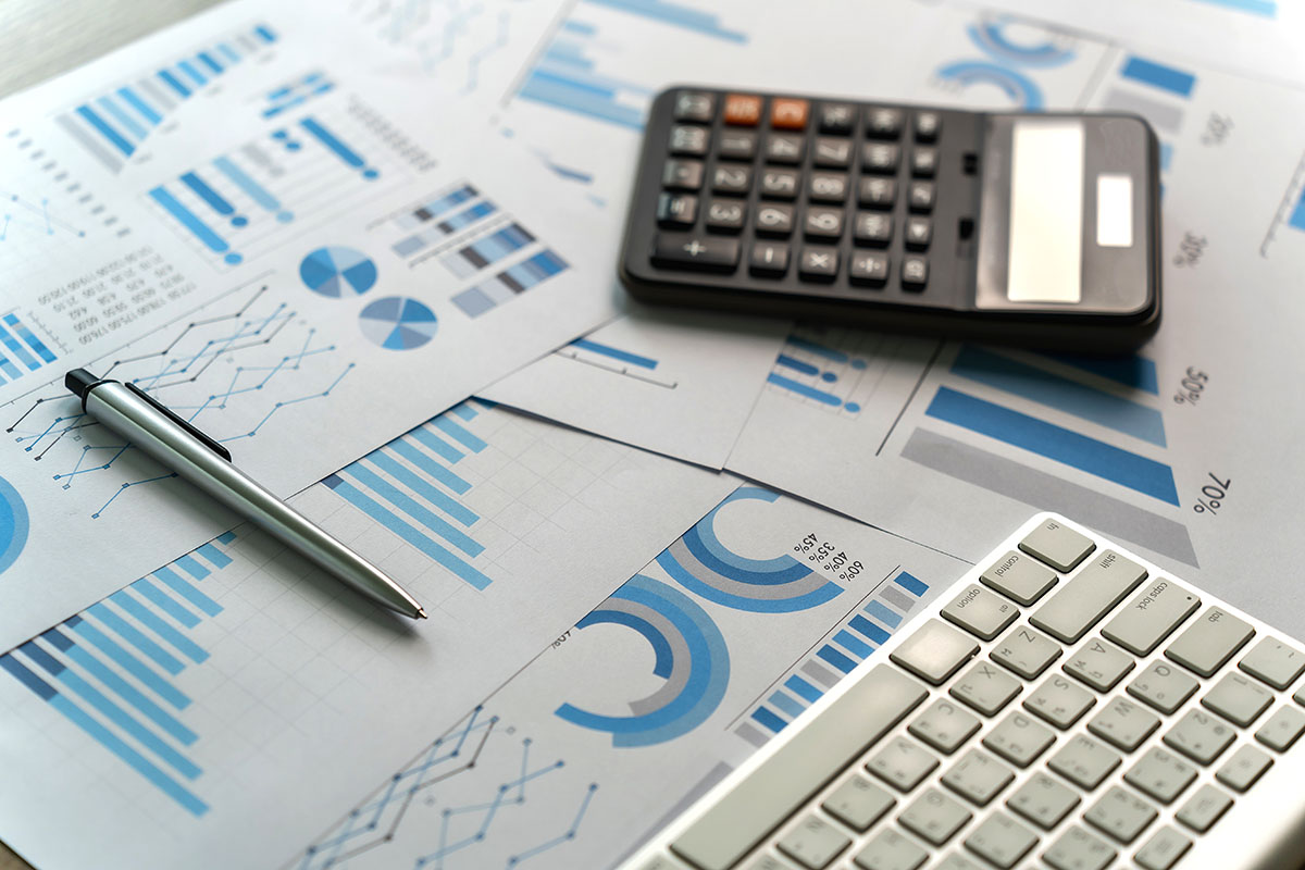 Printed financial reports with bar charts and pie charts spread across a desk, with a calculator, a silver pen and a keyboard