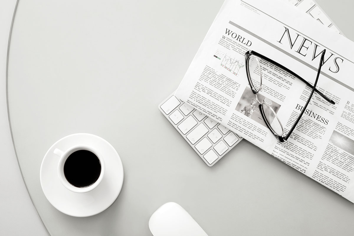 Overhead view of a folded newspaper with World, News and Business headlines and a pair of black reading glasses resting on a white keyboard, beside an espresso cup and a white mouse on a light grey desk