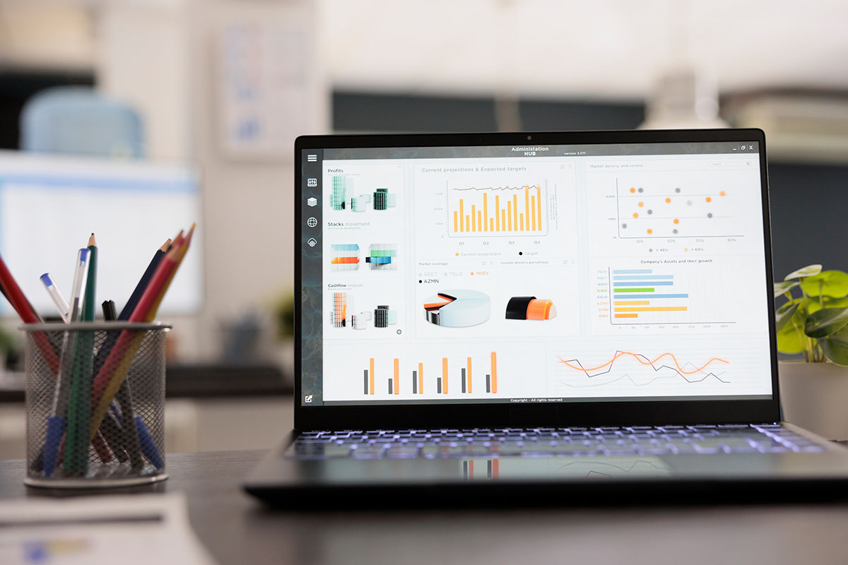 A laptop showing a finance analytics dashboard with bar charts and projections on a desk beside a pencil cup, with a spreadsheet on a monitor in the blurred office behind