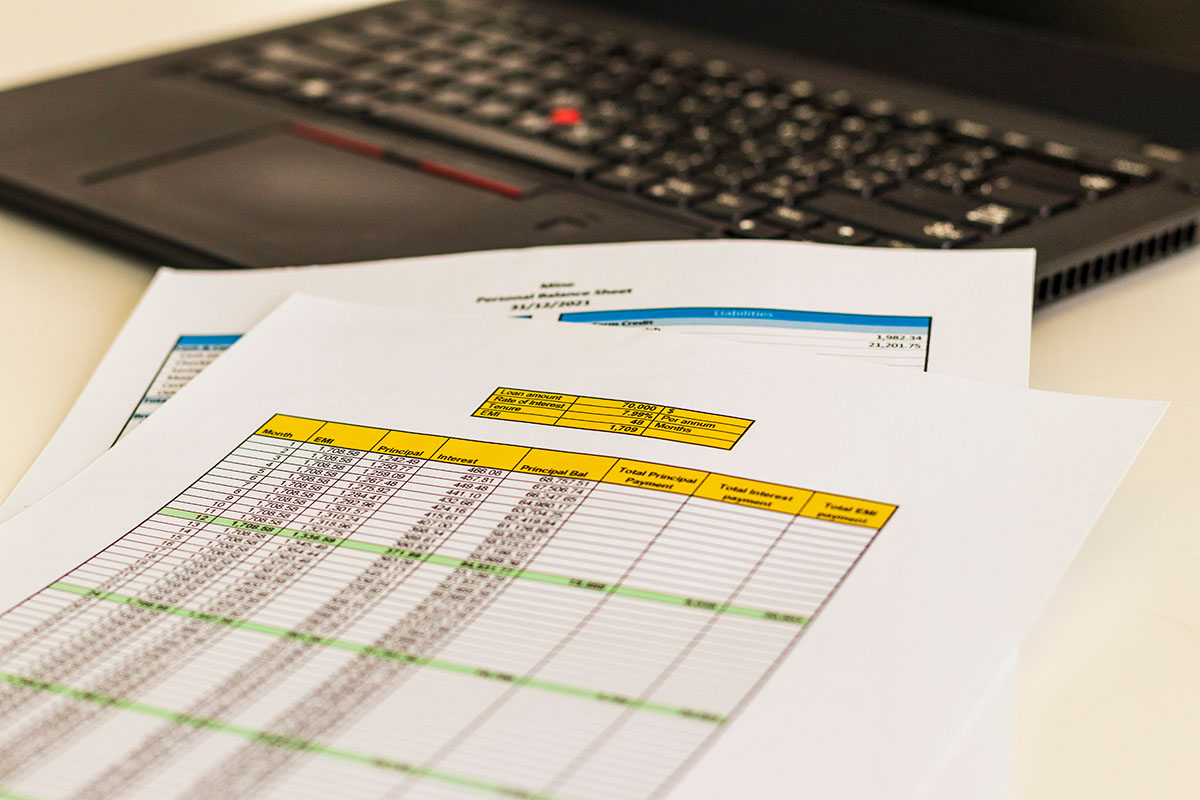 Printed spreadsheet pages with yellow header rows and long columns of payment figures lying on a white desk in front of a black laptop keyboard