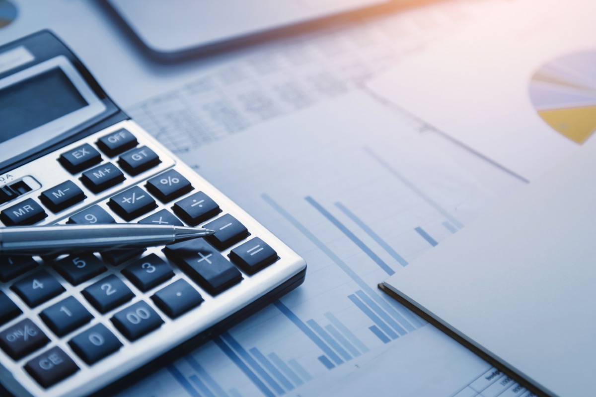 A silver calculator and pen resting on printed bar charts and pie charts on a desk, with a tablet at the top edge of the frame