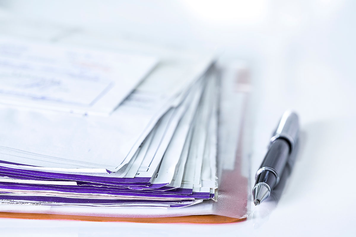 A thick stack of paper invoices and envelopes with purple edges on a white desk beside a silver ballpoint pen