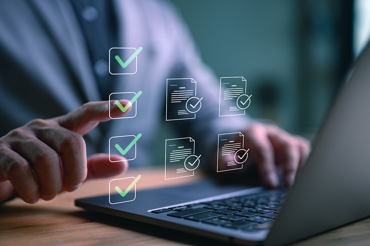 A hand pointing at a column of green digital checkmarks and approved document icons floating above a laptop keyboard on a wooden desk