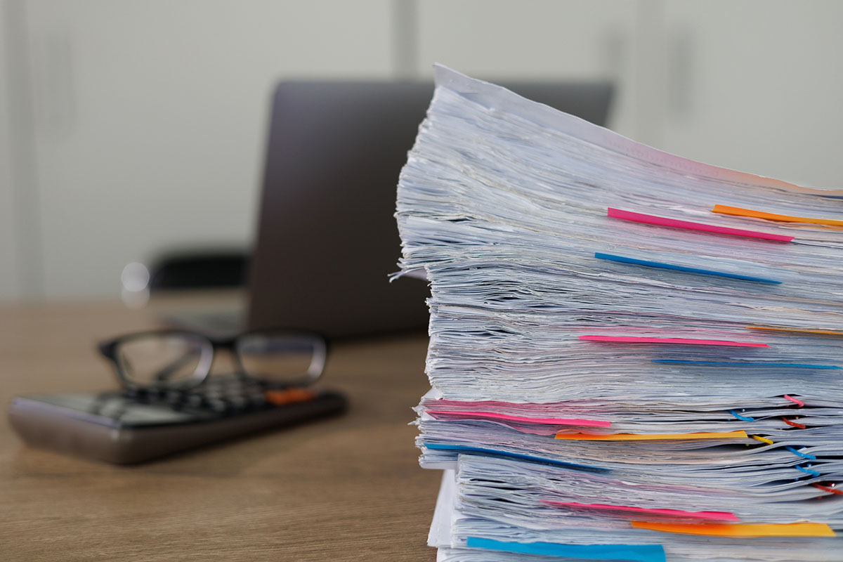 A tall stack of paper documents marked with pink, orange and blue sticky flags on a wooden desk, with reading glasses on a calculator and a laptop blurred in the background