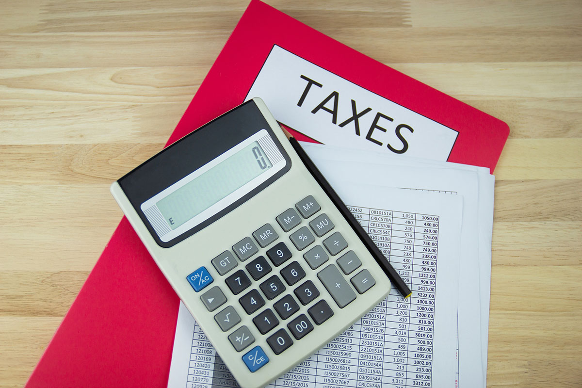 A calculator and pencil resting on printed tables of figures on top of a red folder labeled Taxes, on a light wooden desk