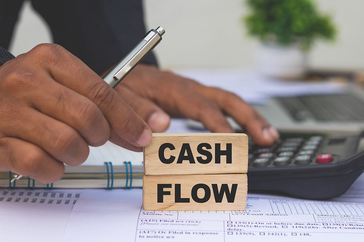 Two wooden blocks spelling Cash Flow on a printed form, with a hand holding a silver pen and another hand on a calculator in the background