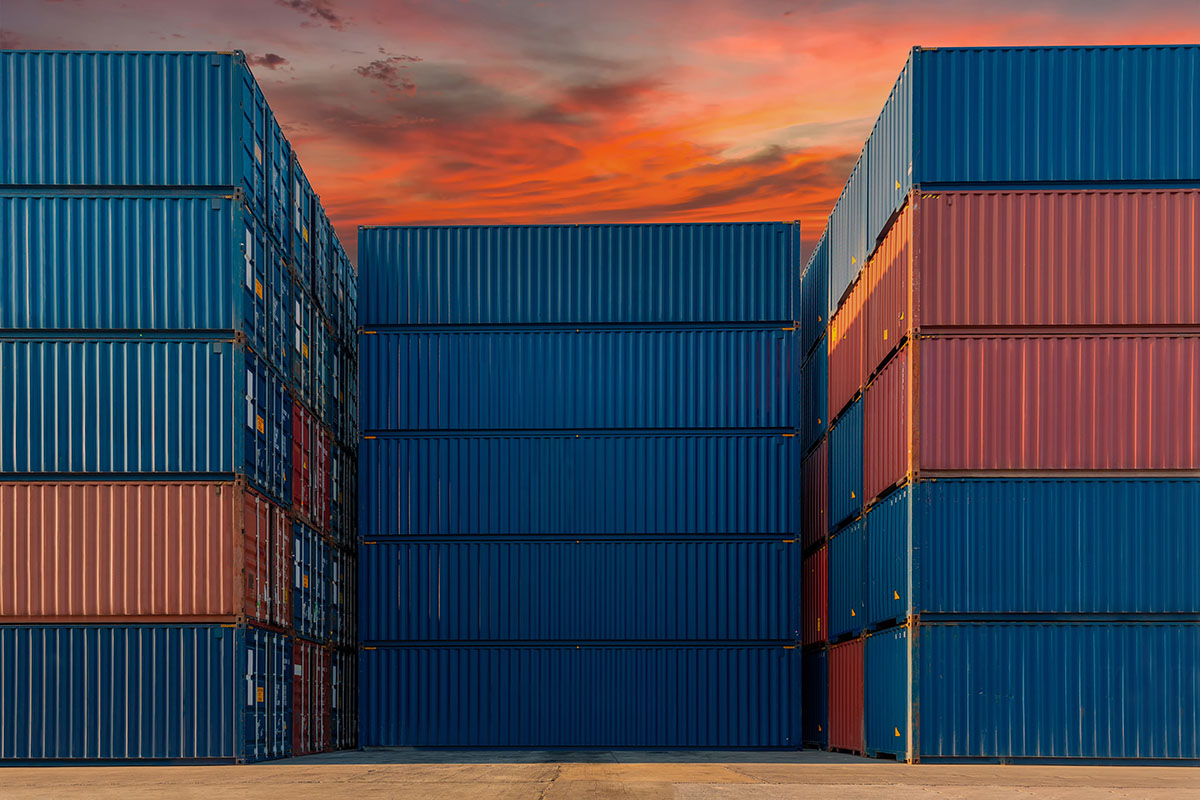 Towering stacks of blue and red shipping containers in a port yard under an orange sunset sky