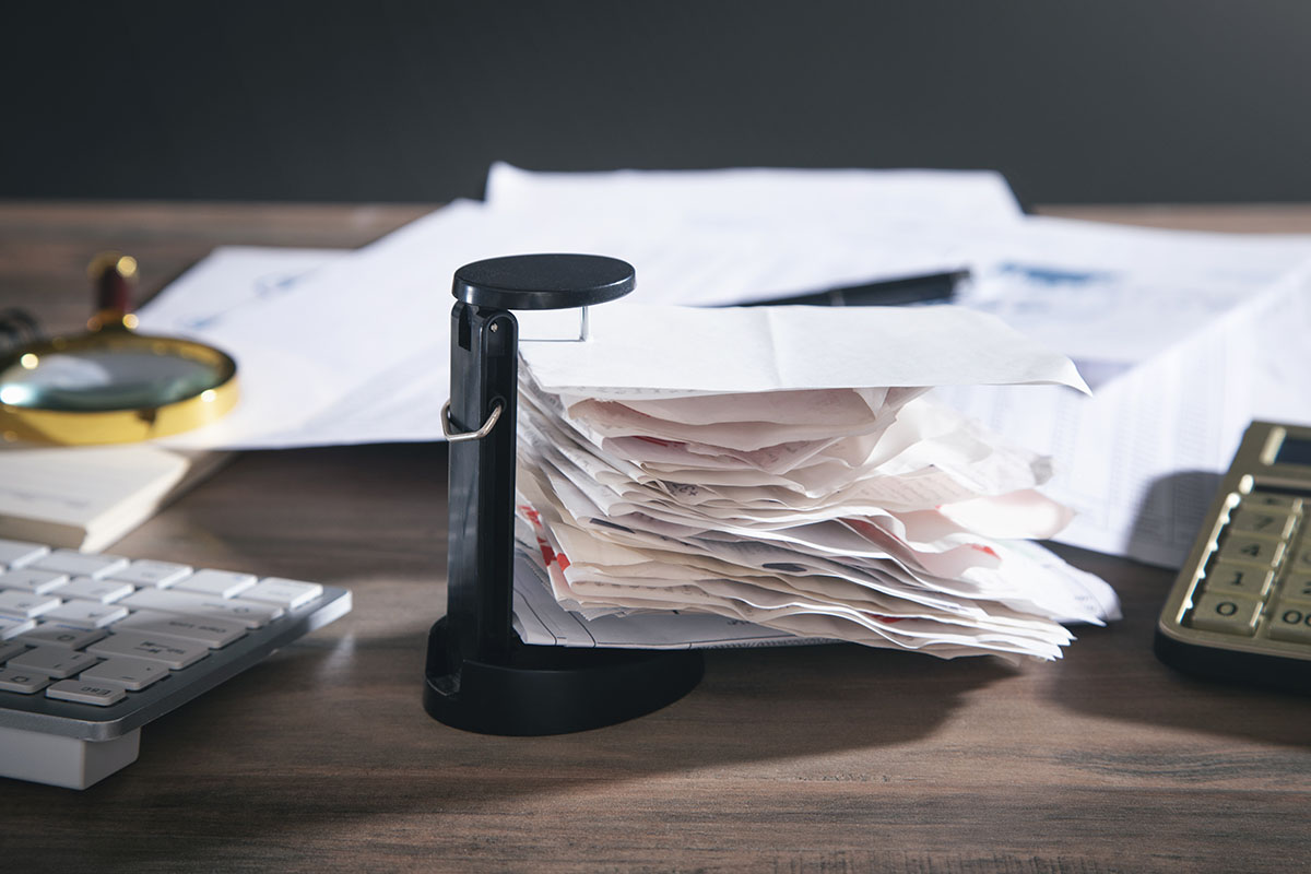 A thick stack of paper bills and receipts held on a black desk spike, with a calculator, keyboard, magnifying glass and financial reports on a wooden desk