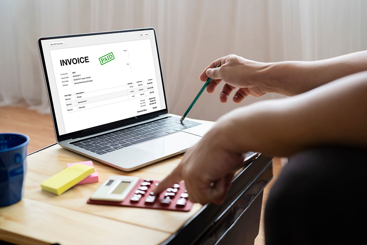 A laptop on a wooden desk showing an invoice stamped paid, while one hand points with a green pencil and the other taps a red calculator beside yellow and pink sticky notes